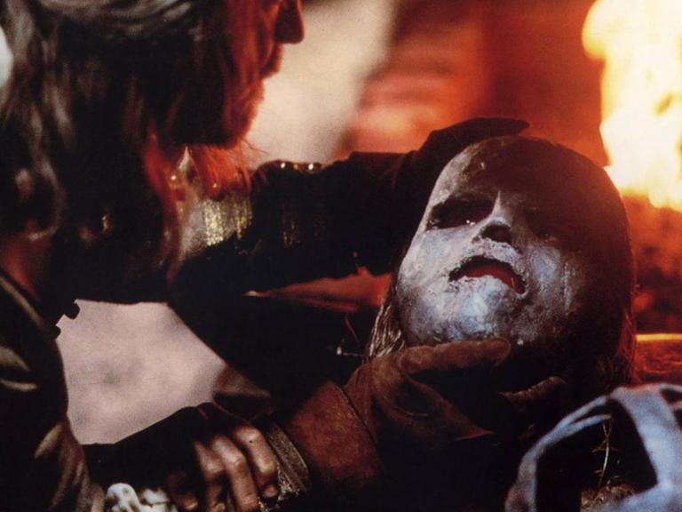 Ein Mann hält eine eiserne Maske über das Gesicht eines anderen Mannes. Dahiner ist ein Feuer zu sehen.