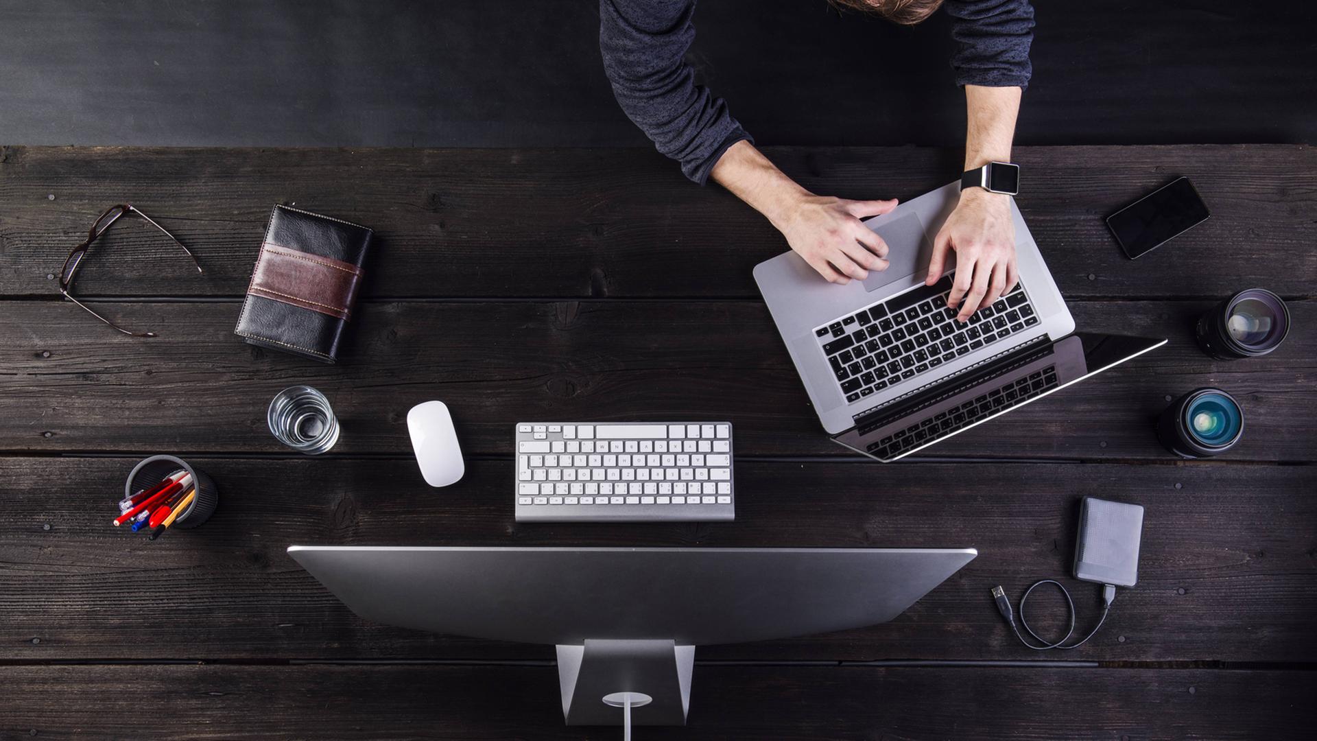 Ein Mann ist von oben zu sehen, wie er an einem Tisch mit dem Computer und mehreren anderen digitalen Geräten wie Notebook, Smartphone, Smartwatch arbeitet.