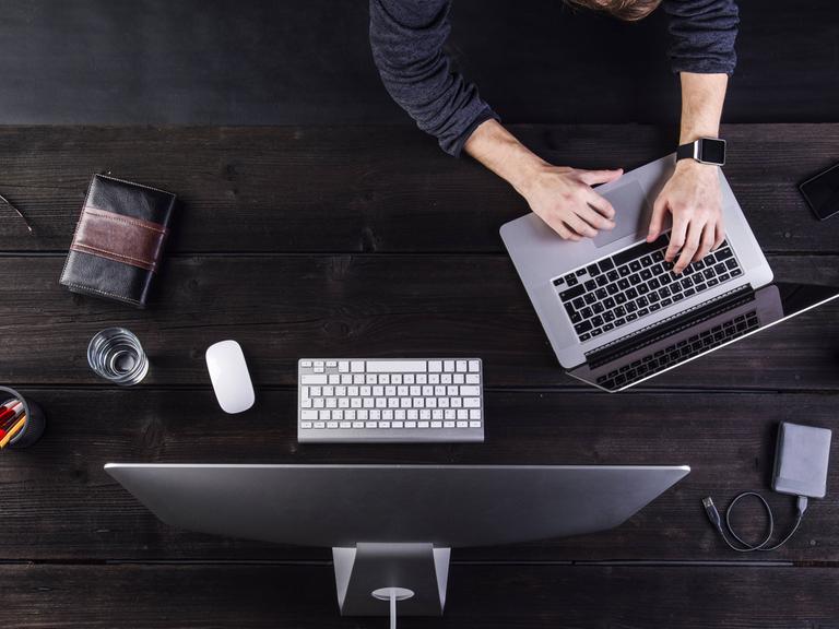 Ein Mann ist von oben zu sehen, wie er an einem Tisch mit dem Computer und mehreren anderen digitalen Geräten wie Notebook, Smartphone, Smartwatch arbeitet.