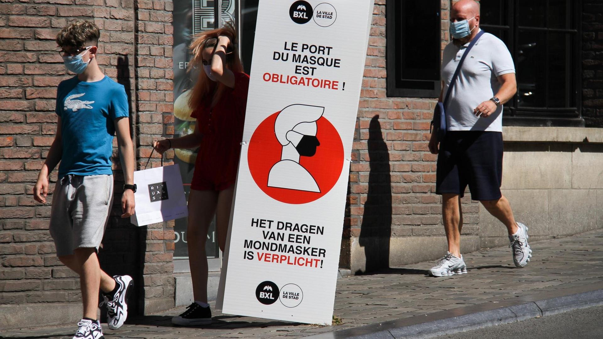 Menschen mit Masken gehten über eine Straße in Brüssel. Dazwischen steht ein Schild, das auf die Maskenpflicht hinweist.