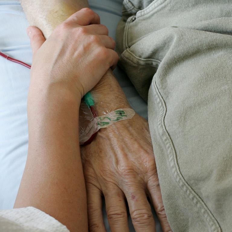 Ein jüngerer Mensch umfasst das Armgelenk einer älteren Person, die im Krankenbett liegt.