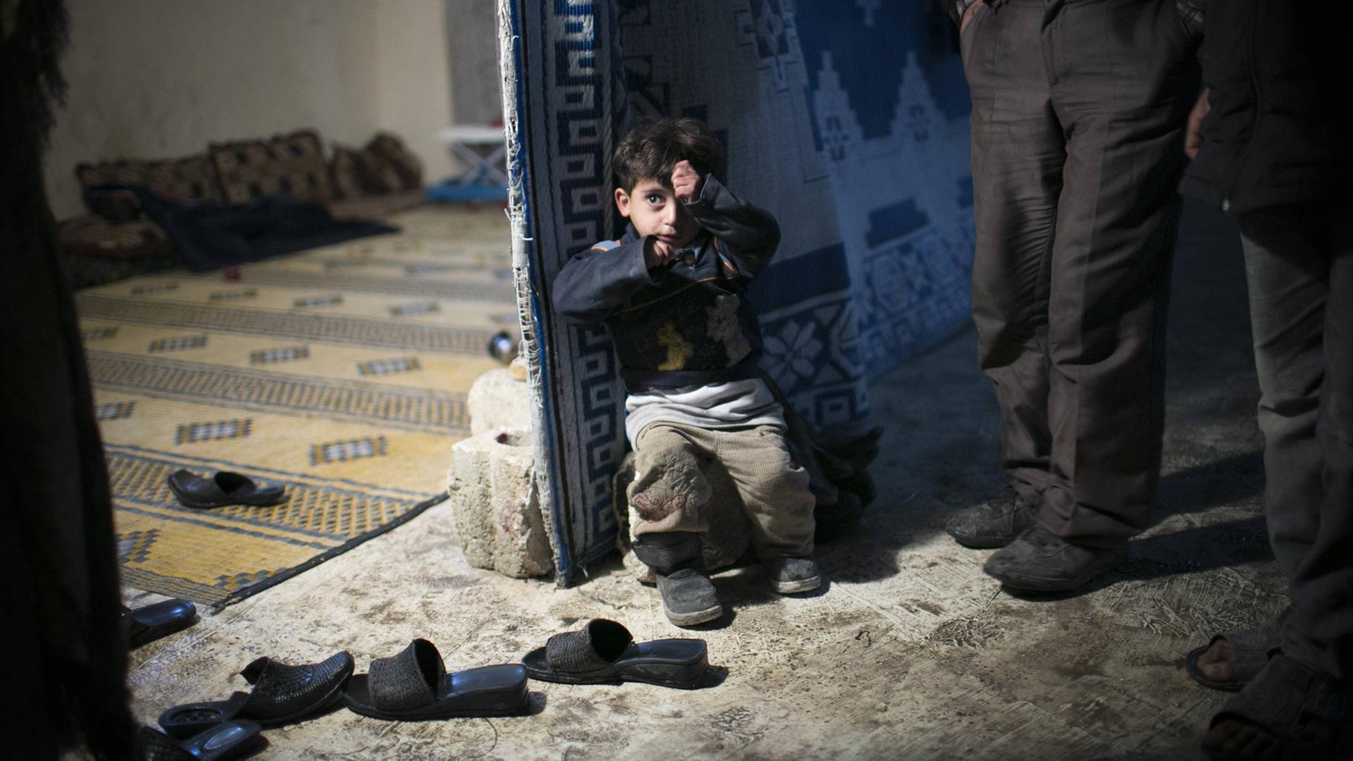 Ein kleiner, ängstlicher, syrischer Junge steht halb in der Knie in einem verlassenen Haus in Aleppo in Syrien. Viele Familien mussten ihr Zuhause verlassen, weil sie entweder in der Nähe von einem militärischen Flugstützpunkt wohnten oder weil ihre Häuser zerbombt wurden.