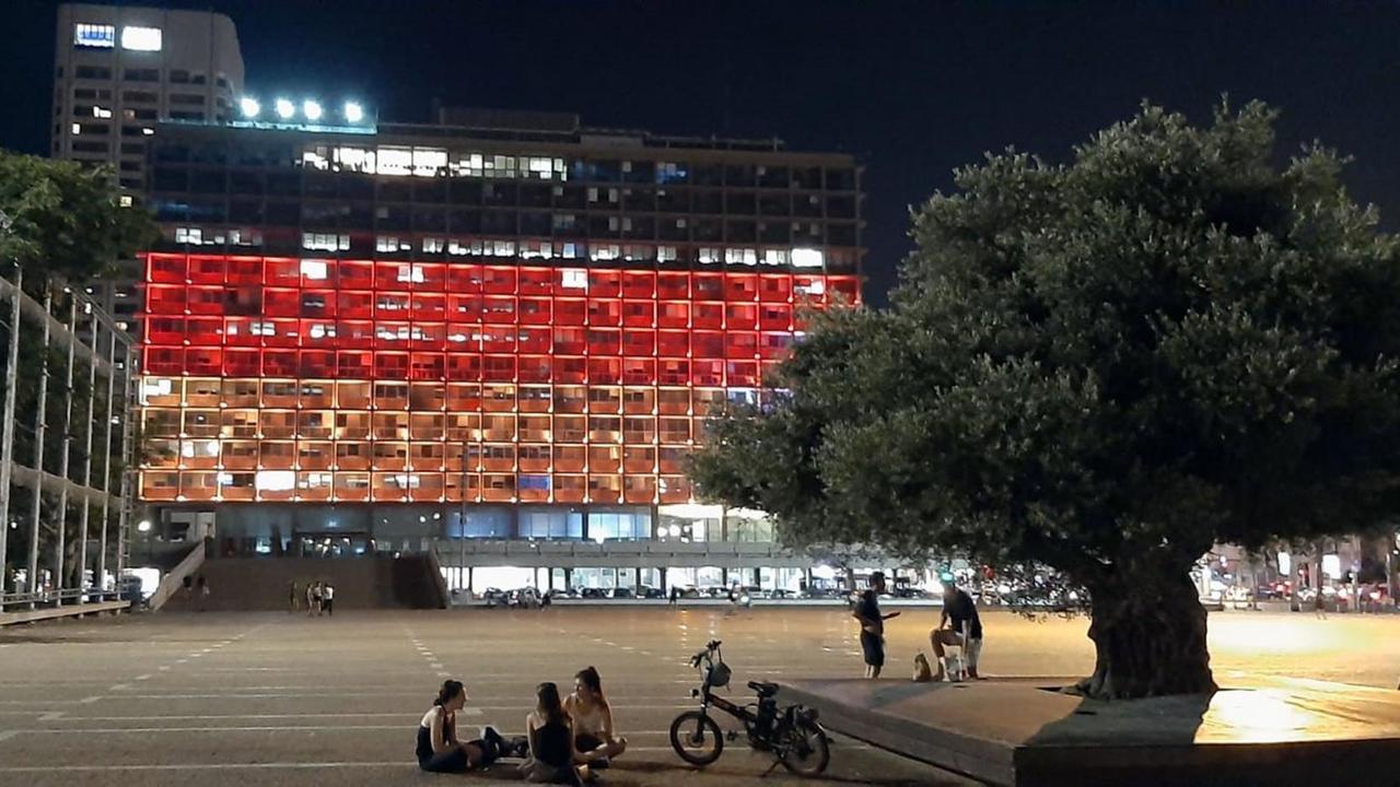 Blick in der Nacht auf das Rathaus, dessen Stockwerke von oben nach unten schwarz-rot-gold beleuchtet sind. Davor ein leerer Parkplatz mit einigen Menschen.