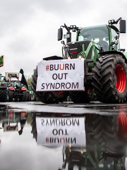 An einem Traktor spiegelt sich in einer Pfütze ein Transparent mit der Aufschrift: "#Buurn out Syndrom".