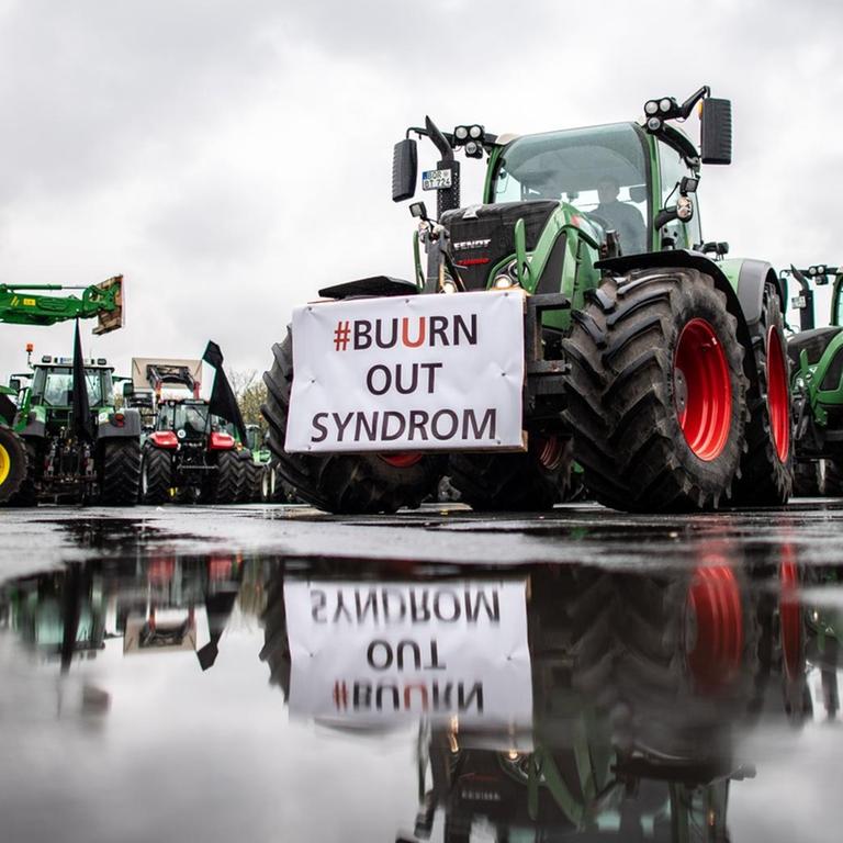 An einem Traktor spiegelt sich in einer Pfütze ein Transparent mit der Aufschrift: "#Buurn out Syndrom". 