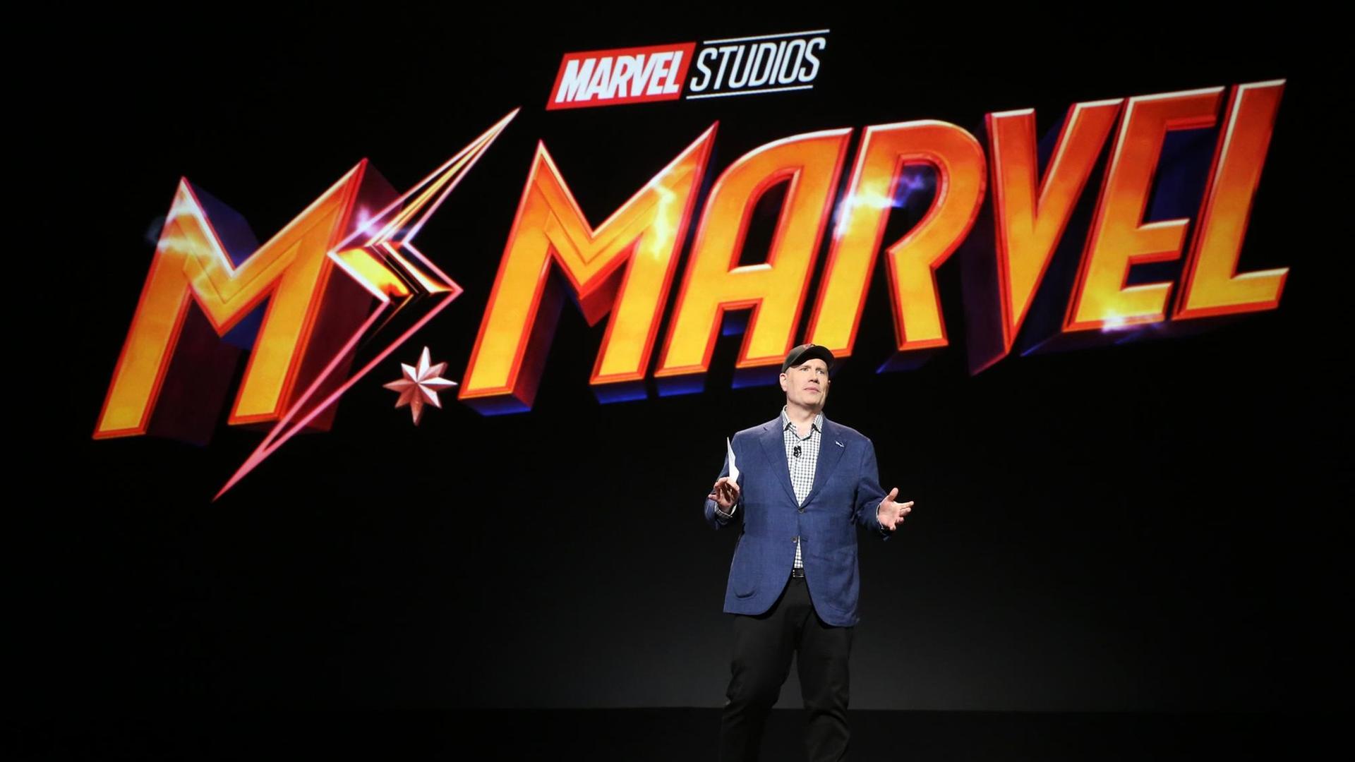 Der Präsident der Marvel Studios, Kevin Feige, auf einer Bühne bei einer Präsentation. Hinter ihm steht in grosser orangener Schrift: Mr. Marvel.
