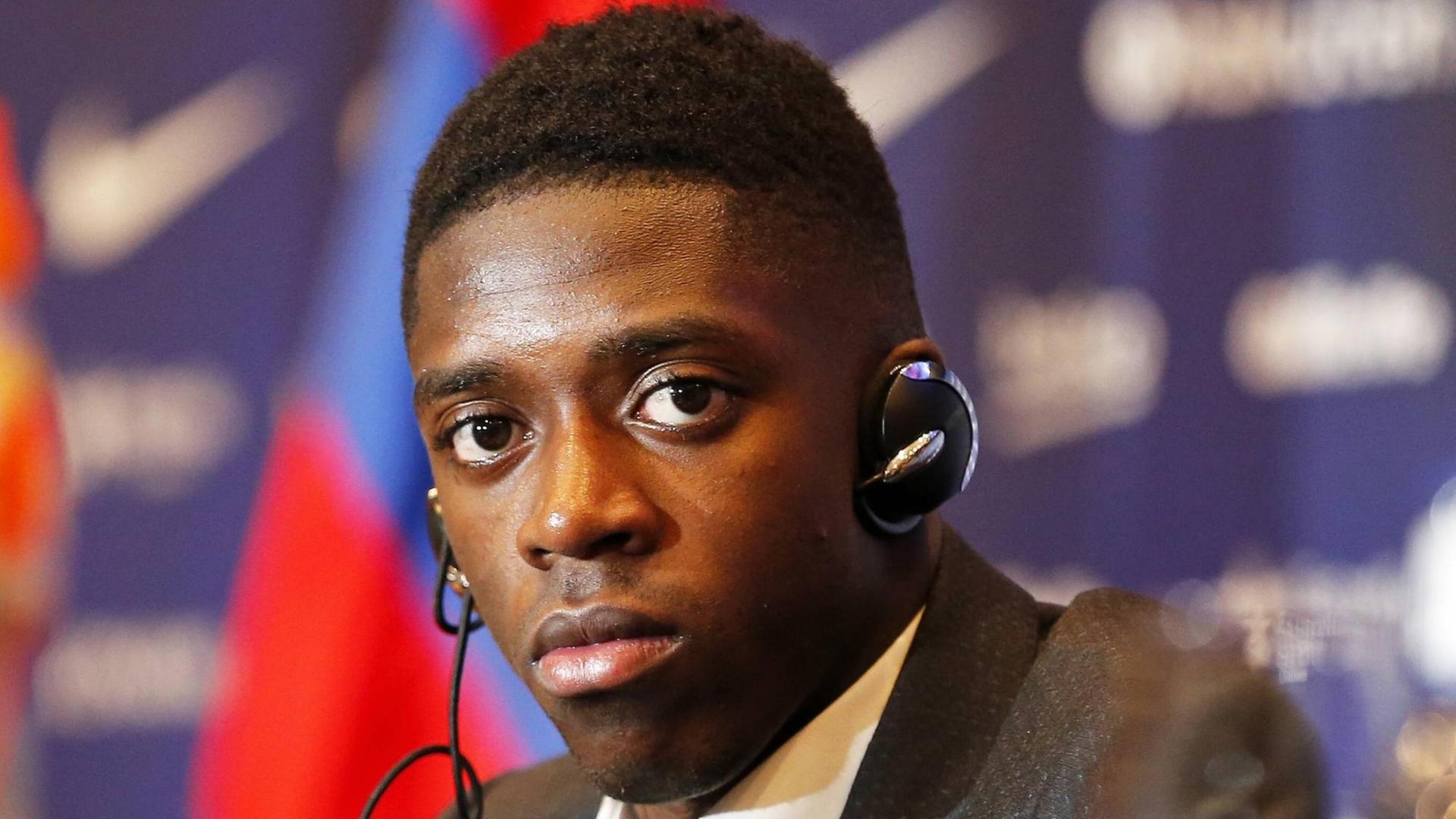 Dembélé sitzt im Anzug und mit Kopfhörern vor einem unscharfen Hintergrund mit den Farben Barcelonas. Er blickt ernst.