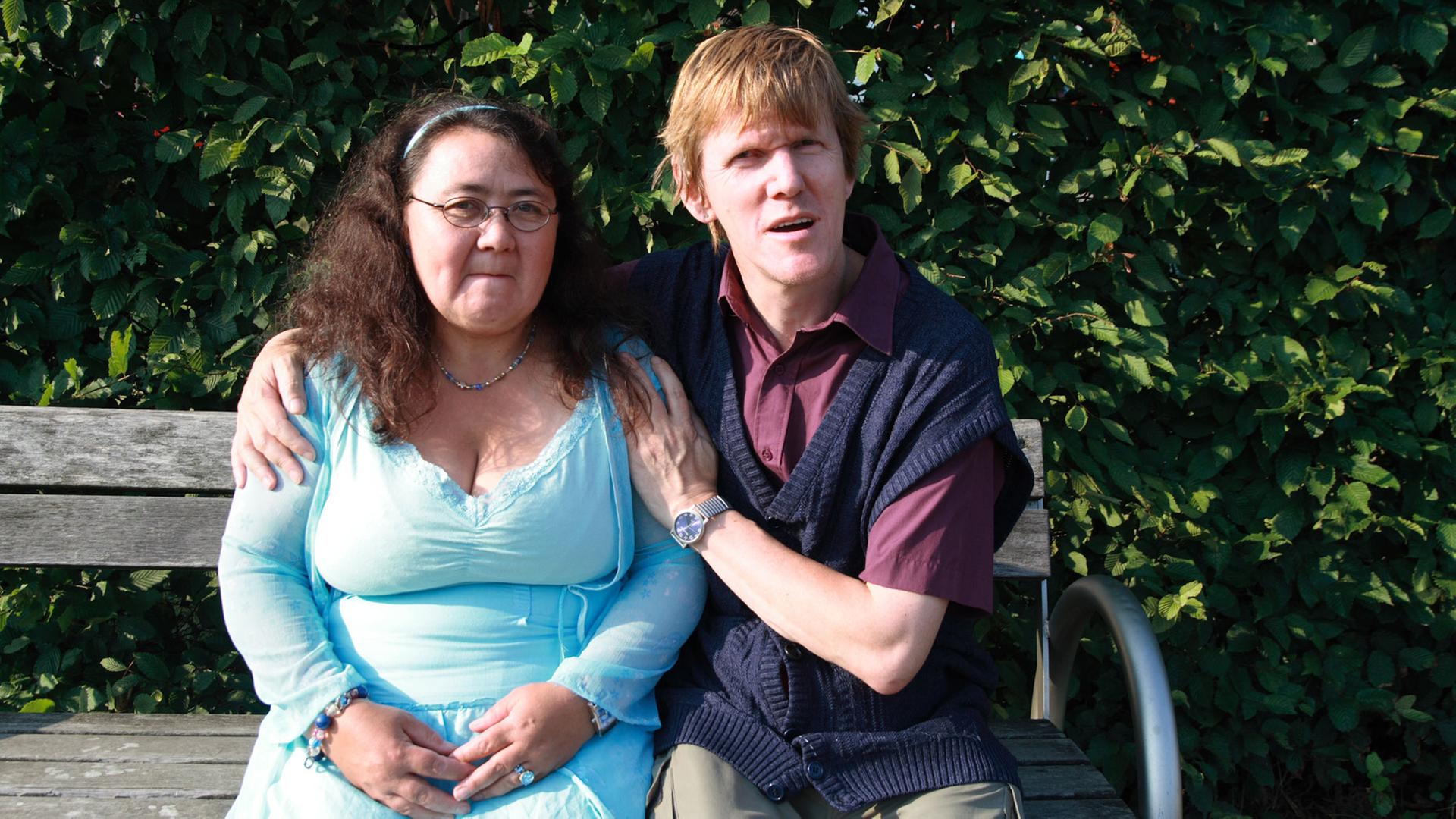 Elsa und Olaf lernten sich über eine Partnervermittlung für Menschen mit Behinderung kennen. Ein junges Paar sitzt auf einer Bank und schaut in die Kamera. Im Hintergrund Bäume von einem Park