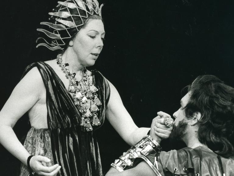 Schwarz-weiß Foto, links im Bild die Sängerin als Dido in einem antiken Kostüm mit Flammen artigen Haarschmuck, rechts knieend der Sänger mit längerem Haar und Bart und Goldschmuck am Arm seine linke Hand hält ihre rechte Hand