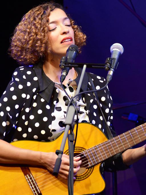 Eine Frau spielt Akustikgitarre und steht mit geschlossenen Augen vor einem Mikrofonständer.