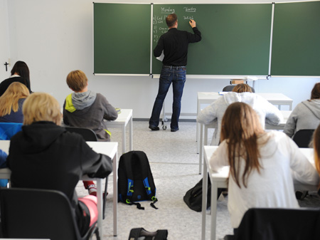 Blick in eine Schulklasse von hinten, auf Rücken von sitzenden Schülern und einen Lehrer an einer Tafel.