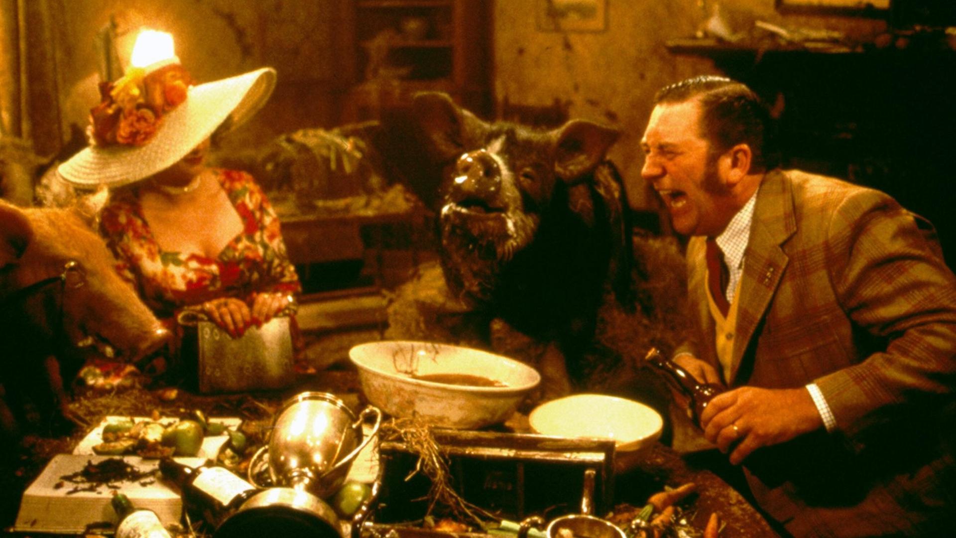 Szene aus dem Film "Animal Farm" von John Stephenson. Eine Frau, ein Schwein und ein Mann sitzen an einem Tisch vollgepackt mit Gegenständen.
