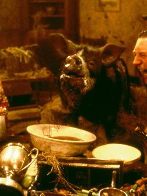 Szene aus dem Film "Animal Farm" von John Stephenson. Eine Frau, ein Schwein und ein Mann sitzen an einem Tisch vollgepackt mit Gegenständen.