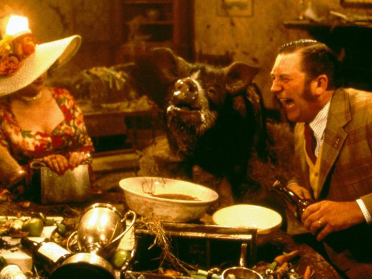 Szene aus dem Film "Animal Farm" von John Stephenson. Eine Frau, ein Schwein und ein Mann sitzen an einem Tisch vollgepackt mit Gegenständen.