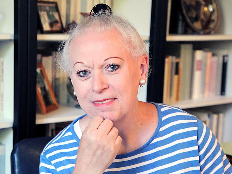 Eine Frau jenseits der 60 mit weißen hochgesteckten Haaren. Die Augenbrauen sind gezupft und mit einem auffällig dunkelbraunen Strich nachgezogen. Augen und Lippen sind geschminkt. Die Person trägt Ohrklipse und ein blau-weißes Ringelshirt.