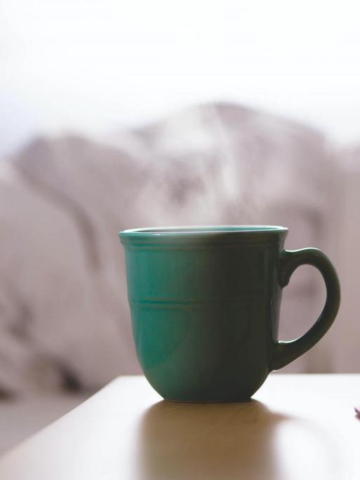 Eine Tasse mit heißem Getränk steht auf einem Tisch neben einem ungemachten Bett.