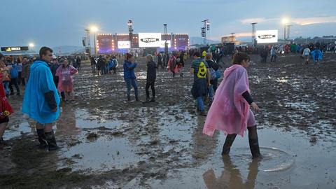 Festivalbesucher mit Regenjacken waten durch den Schlamm, im Hintergrund die beleuchtete Bühne.