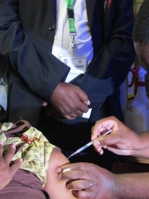 Die Gesundheitsminsterin von Namibia sitzt in einem gelben Kleid auf einem Stuhl und wird von einem Arzt geimpft