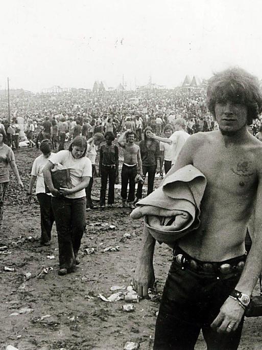 Woodstock 1969, schwarz-weiß Fotografie: Festivalbesucher laufen über den schlammigen Boden. Rechts eine junge Frau mit Sonnenbrille und ein Mann ohne T-Shirt.
