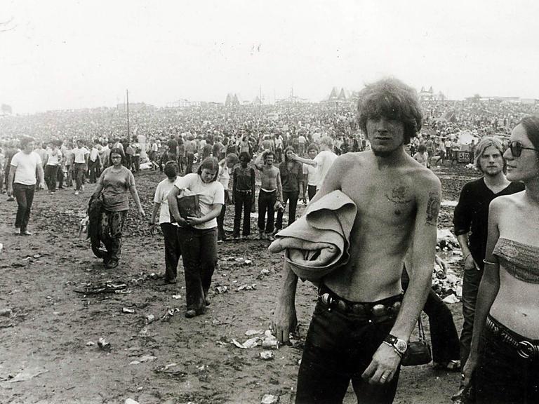 Woodstock 1969, schwarz-weiß Fotografie: Festivalbesucher laufen über den schlammigen Boden. Rechts eine junge Frau mit Sonnenbrille und ein Mann ohne T-Shirt.