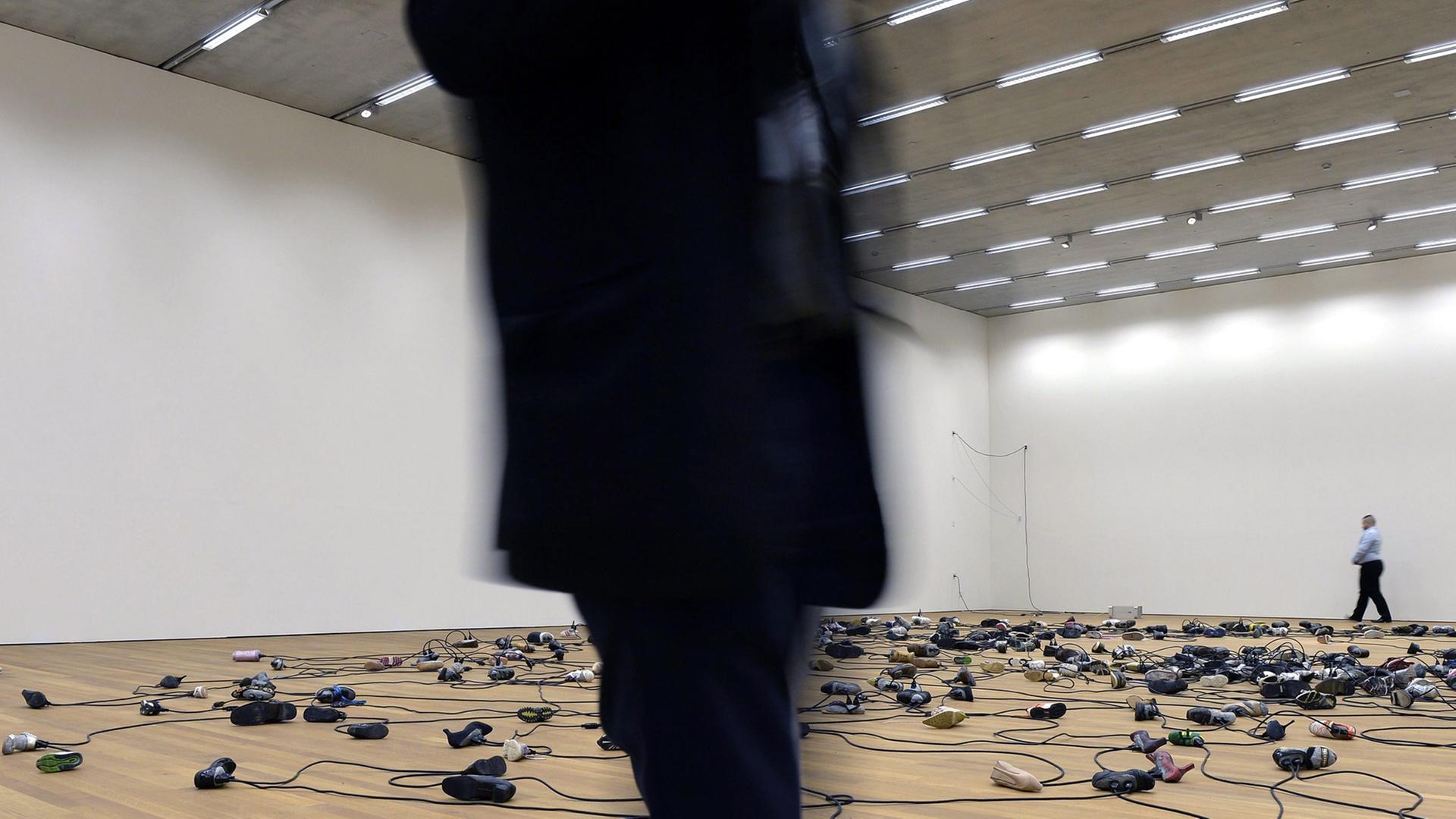 Miteinander verkabelte Schuhe liegen auf dem Boden einer Museumshalle. Ein schwarz gekleideter Mann läuft durchs Bild.