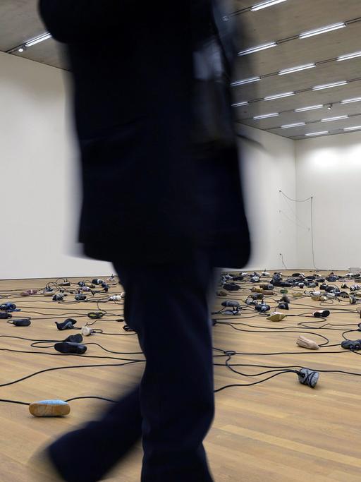 Miteinander verkabelte Schuhe liegen auf dem Boden einer Museumshalle. Ein schwarz gekleideter Mann läuft durchs Bild.