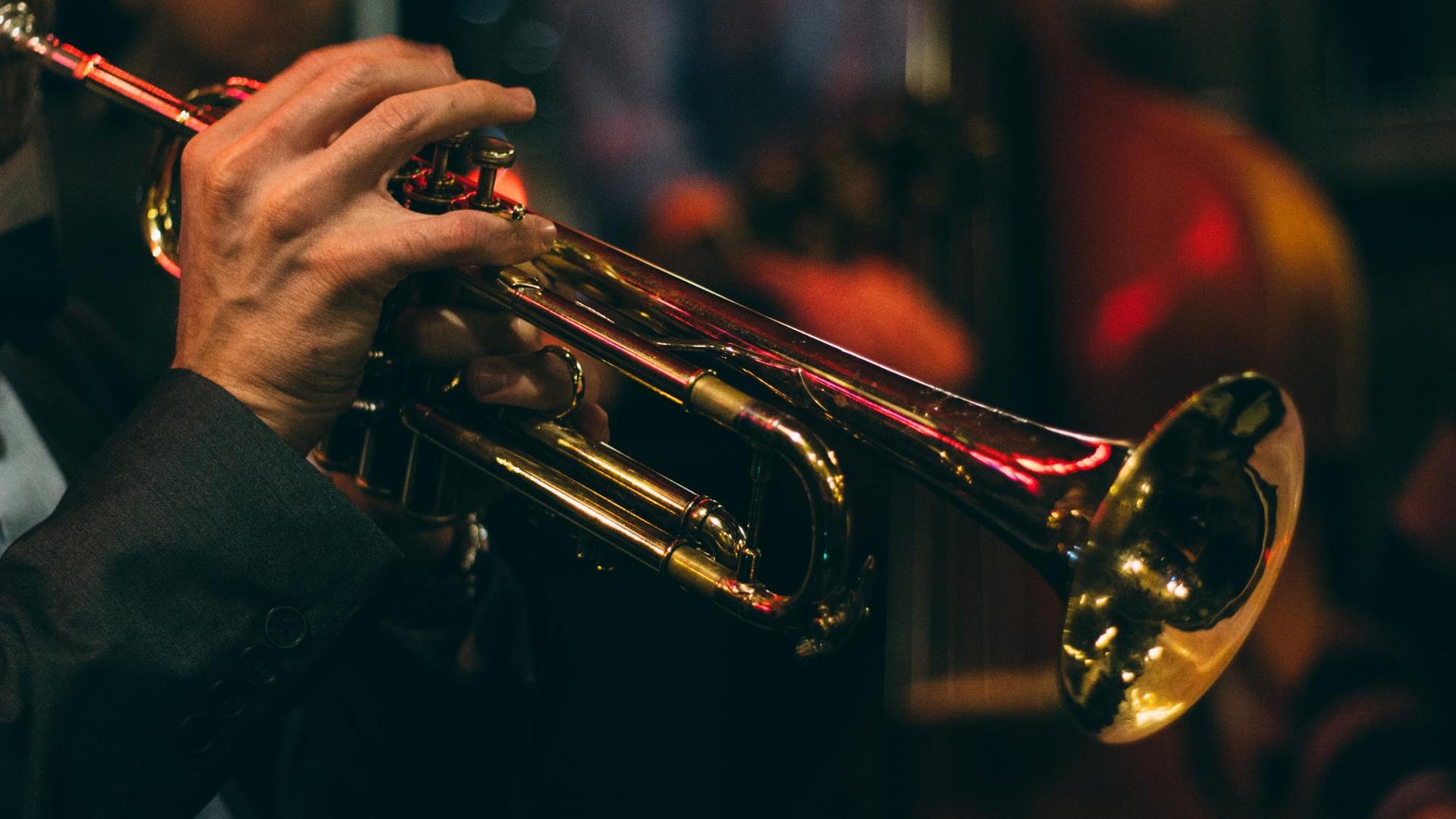 Detail einer Hand die auf einer Jazztrompete spielt. Im Hintergrund das stimmungsvolle Licht einer Bar.
