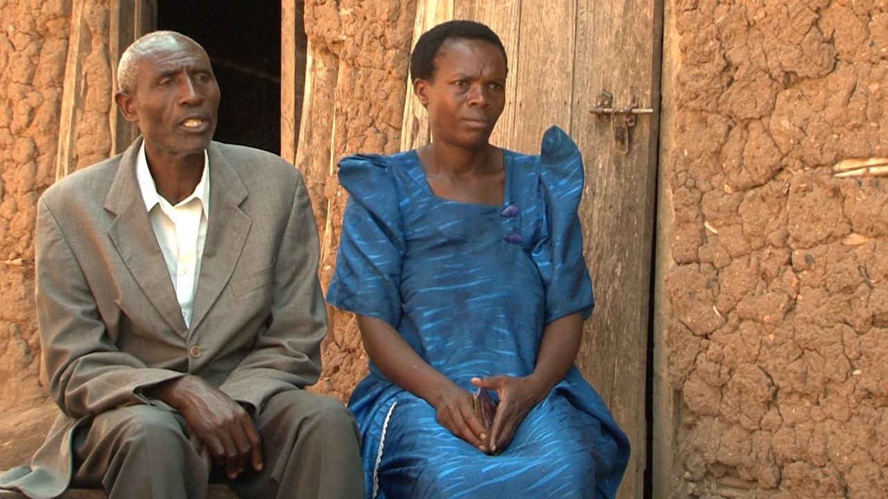 Der ugandische Kleinbauer Aminadabu Kasoma und seine Ehefrau Vanisi sitzen auf einer Holzbank. Sie trägt ein blaues Kleid, er einen braunen Anzug mit Hemd.