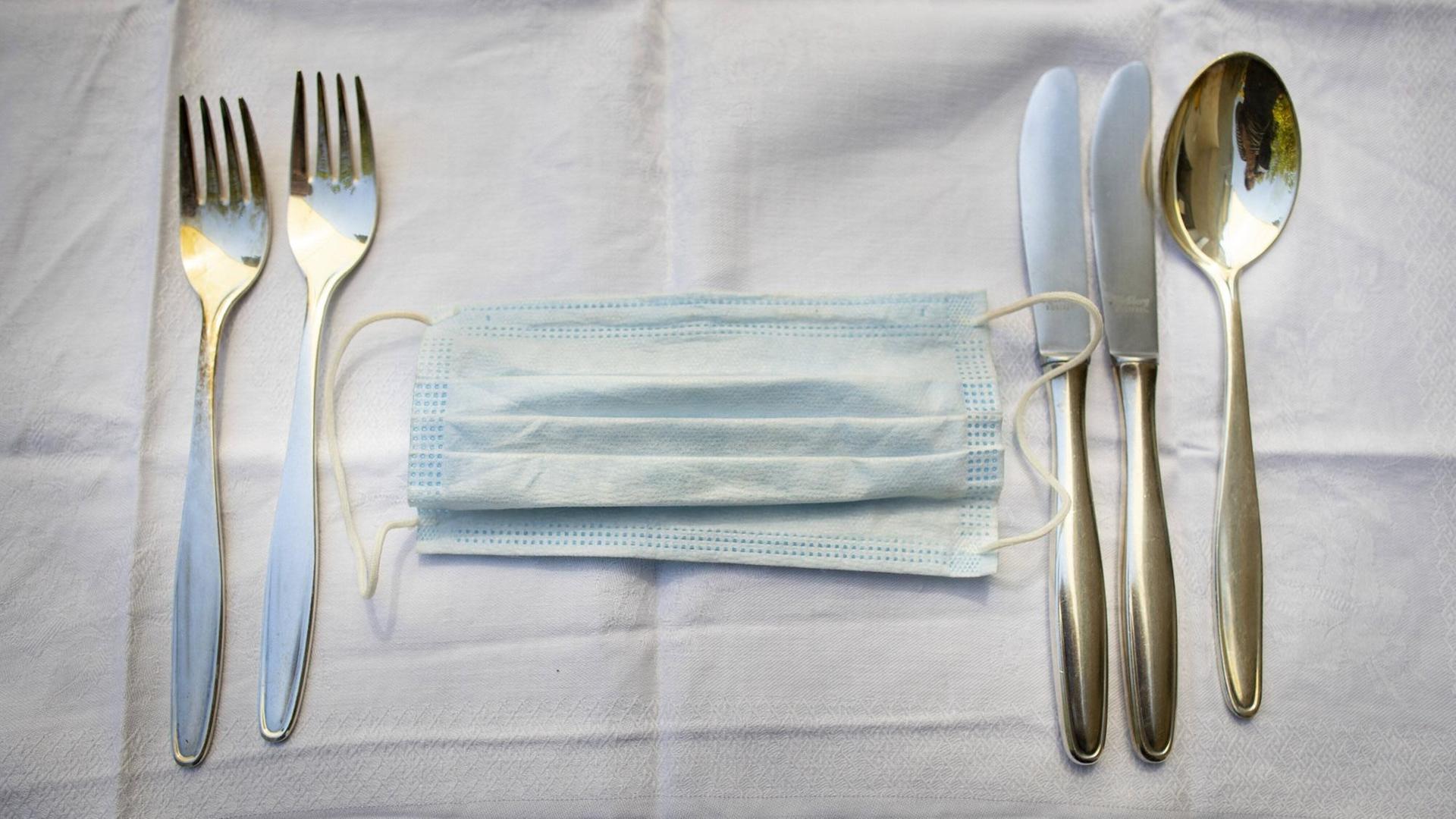 Ein medizinischer Mundschutz zwischen Messer und Gabel auf einer Tischdecke im Restaurant.