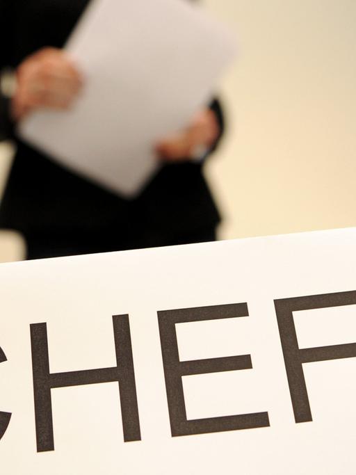 Ein Schild mit der Aufschrift "Chefin". Verschwommen im Hintergrund: Eine Frau in Business-Kleidung, die Papier in Händen hält.