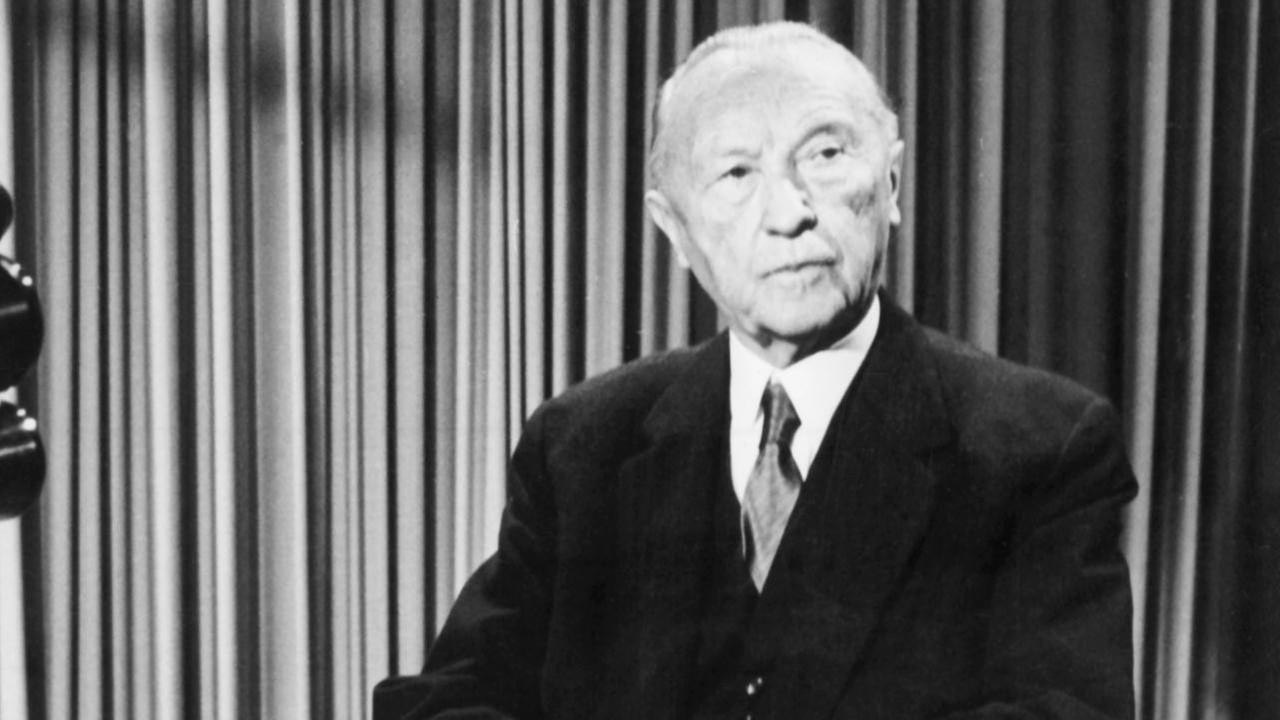 Bundeskanzler Konrad Adenauer sitzt während der Aufzeichnung einer Fernsehansprache in einem Fernsehstudio hinter einem Tisch.