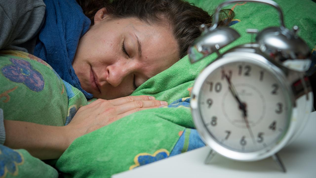 Eine Frau schläft im Bett, während ein Wecker neben ihr auf dem Nachttischränkchen steht.