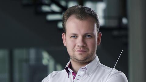 Ein junger Mann im weißen Hemd mit Dirigentenstab in der Hand blickt stolz den Betrachter an.