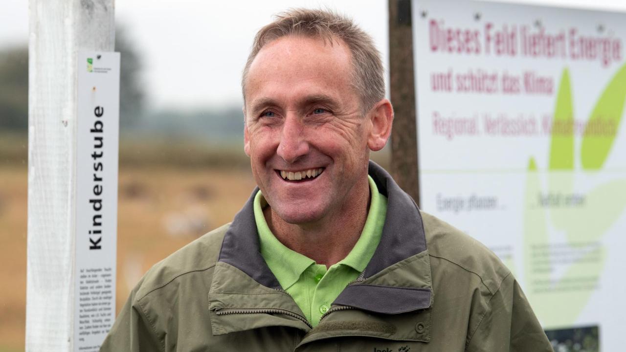 Landwirt Peter Kaim, Inhaber des Havellandhofes Ribbeck, lacht neben einem Schild mit der Aufschrift "Dieses Feld liefert Energie und schützt das Klima". 