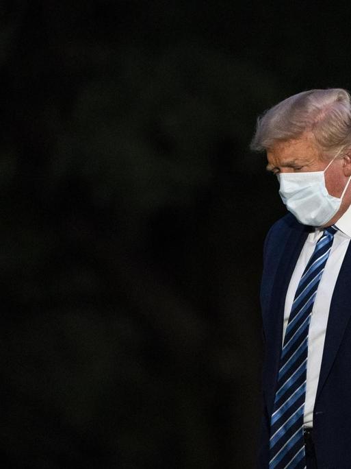 Donald Trump trägt eine Maske und steht mit geneigtem Kopf vor einem schwarzen Hintergrund