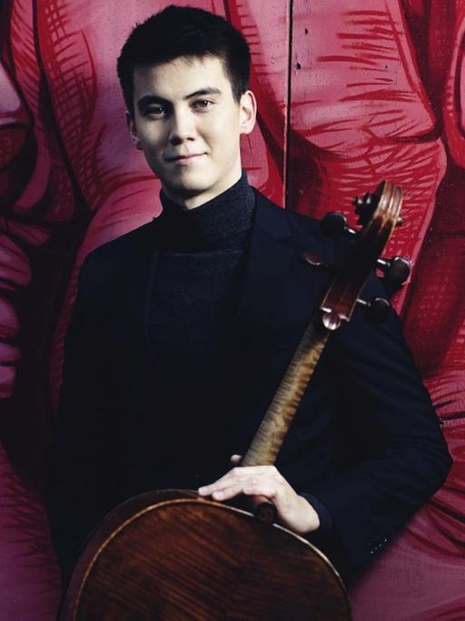 Der junge Cellist mit schwarzen, kurzen Haaren steht mit seinem Instrument vor einem Graffiti, das eine große, rote Hand mit gekrümmten Fingern zeigt.