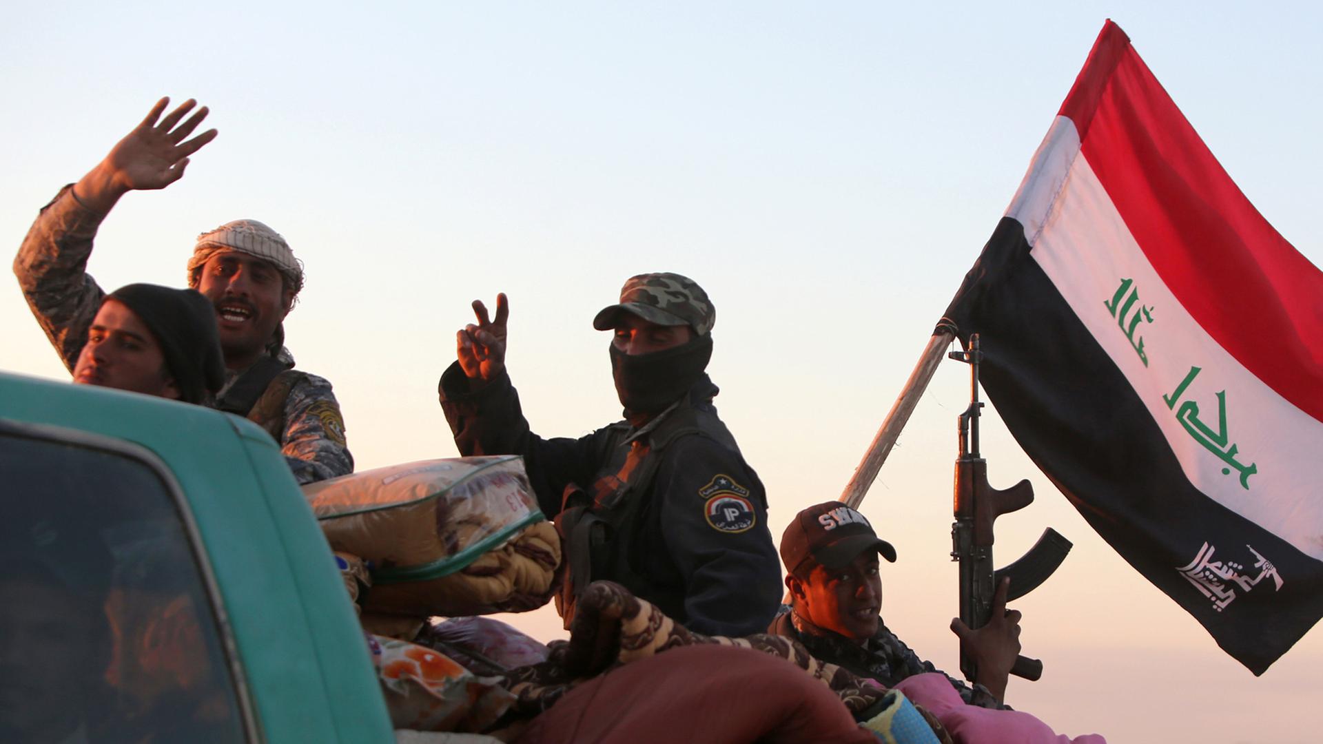 Mehrere Männer sitzen winkend mit einer irakische Flagge auf der offenen Ladefläche eines Pick-up-Trucks.