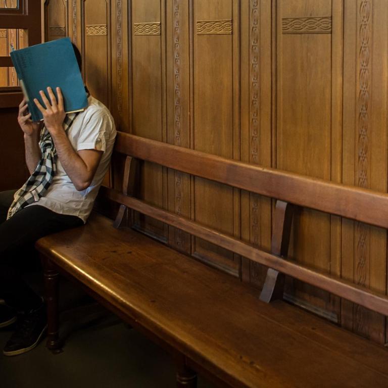 Ein Angeklageter sitzt auf einer langen hölzernen Anklagebank am Langericht Berlin-Moabit. Er verdeckt sein Gesicht mit einem Aktendeckel.
