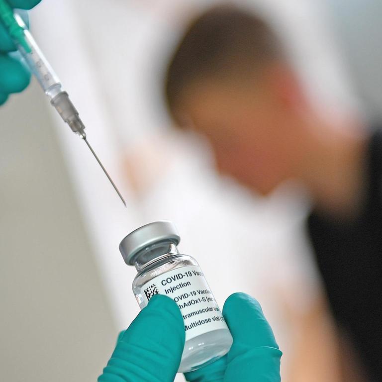 Mit einer Spritze wird Impfserum gegen Covid 19 aus einer Ampulle gezogen. Im Hintergrund ist ein Jugendlicher zu erkennen, der geimpft werden soll.