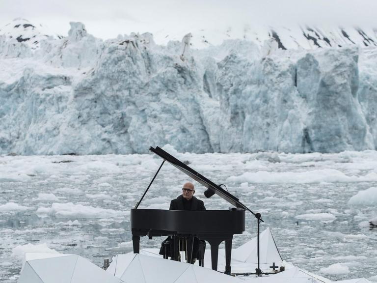Ludovico Einaudi sitzt an einem Flügel, der auf einer Plattform im eisschollendurchsetzten Meer schwimmt, im Hintergrund Eisberge