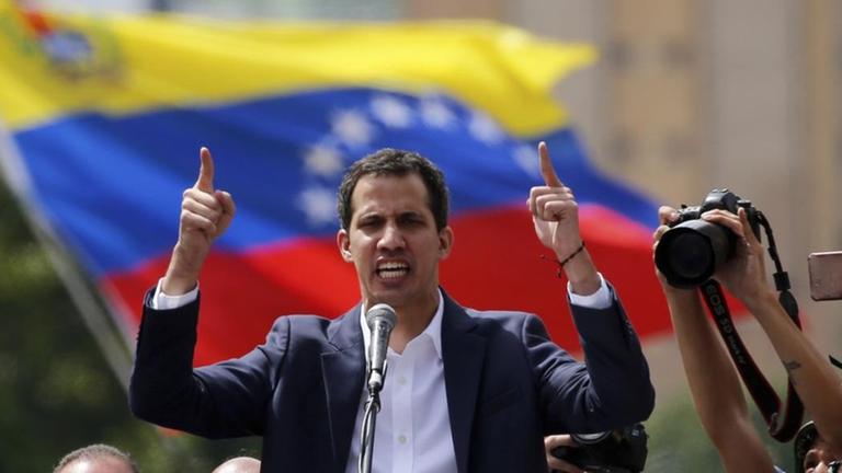 Ein Mann spricht an einem Rednerpult, um ihn herum sind Flaggen des Landes Venezuela. 

