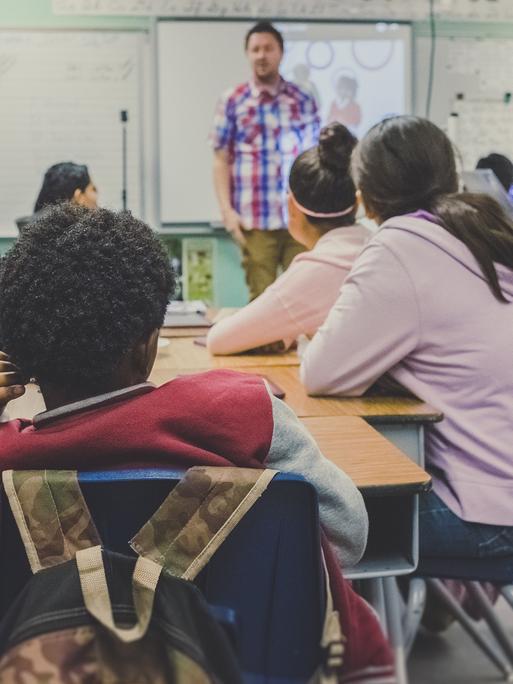 Ein Schülergruppe sitzt während des Unterrichts an einem Tisch - vorn steht der Lehrer und erklärt etwas.