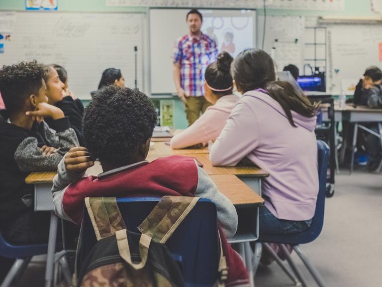 Ein Schülergruppe sitzt während des Unterrichts an einem Tisch - vorn steht der Lehrer und erklärt etwas.