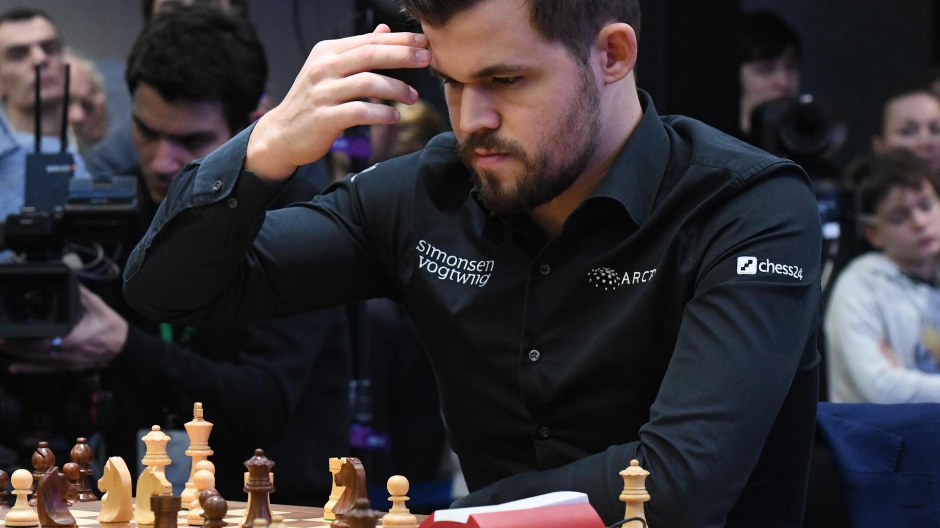Schachweltmeister Magnus Carlsen mit kurzrasiertem Bart und schwarzem Hemd sitz vor einem Schachbrett und hält sich die Hand an den Kopf.