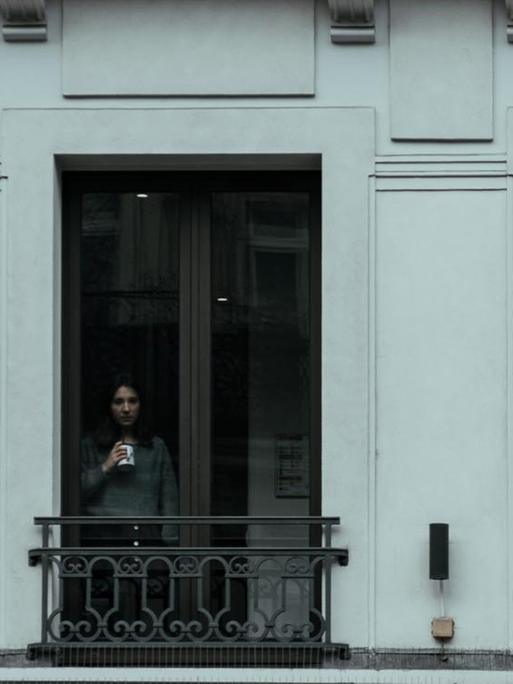 Eine Frau steht mit einer Tasse in der Hand an einem Fenster.