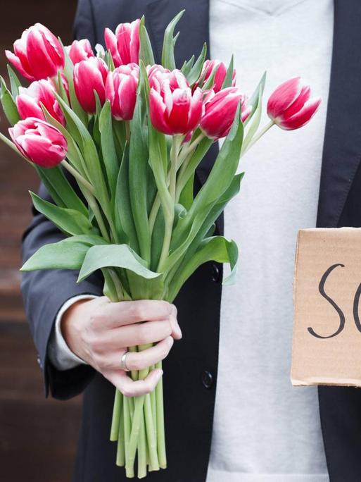 Das Bild zeigt einen Mann, der einen Blumenstrauß und ein Pappschild in den Händen hat. Auf dem Schild steht "sorry".