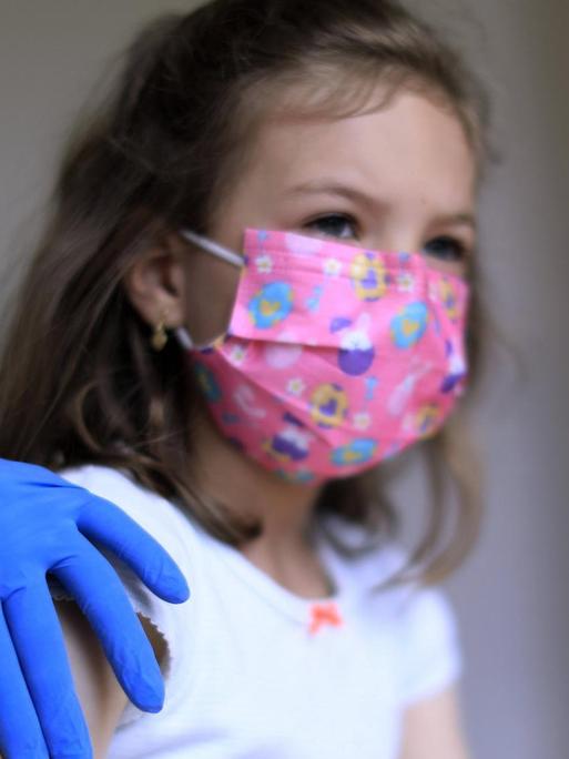 Einem Mädchen mit rosa Mund-Nasenschutz wird mit einer Spritze eine Impfung in den Oberarm injiziert.