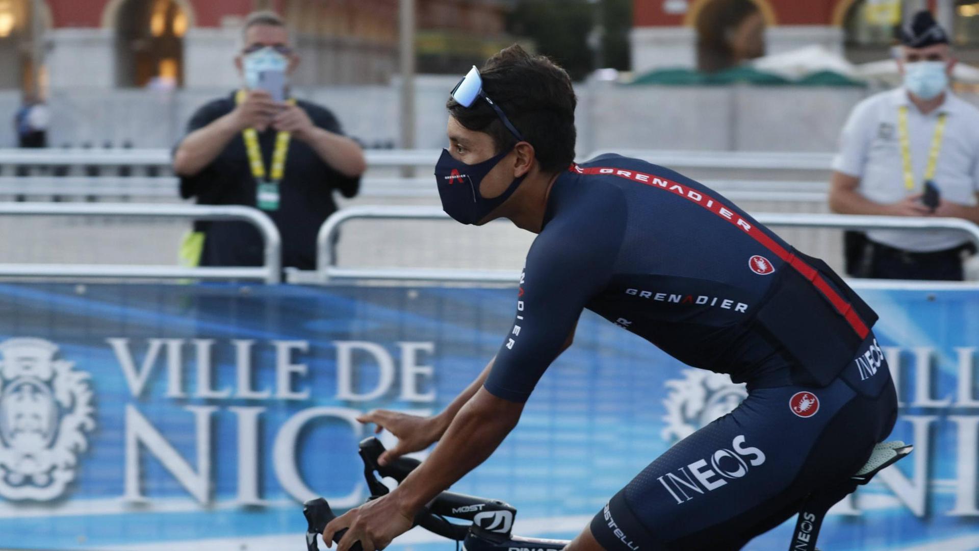 Ein Radfahrer der Tour de France mit Mund-Nase-Schutz fährt in Nizza an einem Absperrgitter vorbei, hinter dem zwei Männer Kamera stehen.