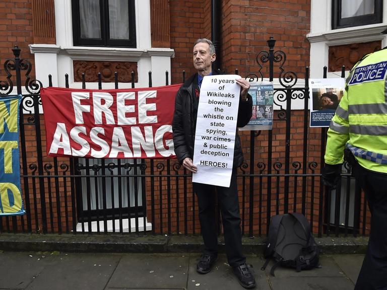 Der Mann steht vor dem Zaun der Botschaft und trägt ein Transparent, auf dem Wikileaks als "Heroes" bezeichnet wird. Ein Polizist läuft an ihm vorbei. Am Zaun hänge Transparente, eines mit der Aufschrift "Free Assange".