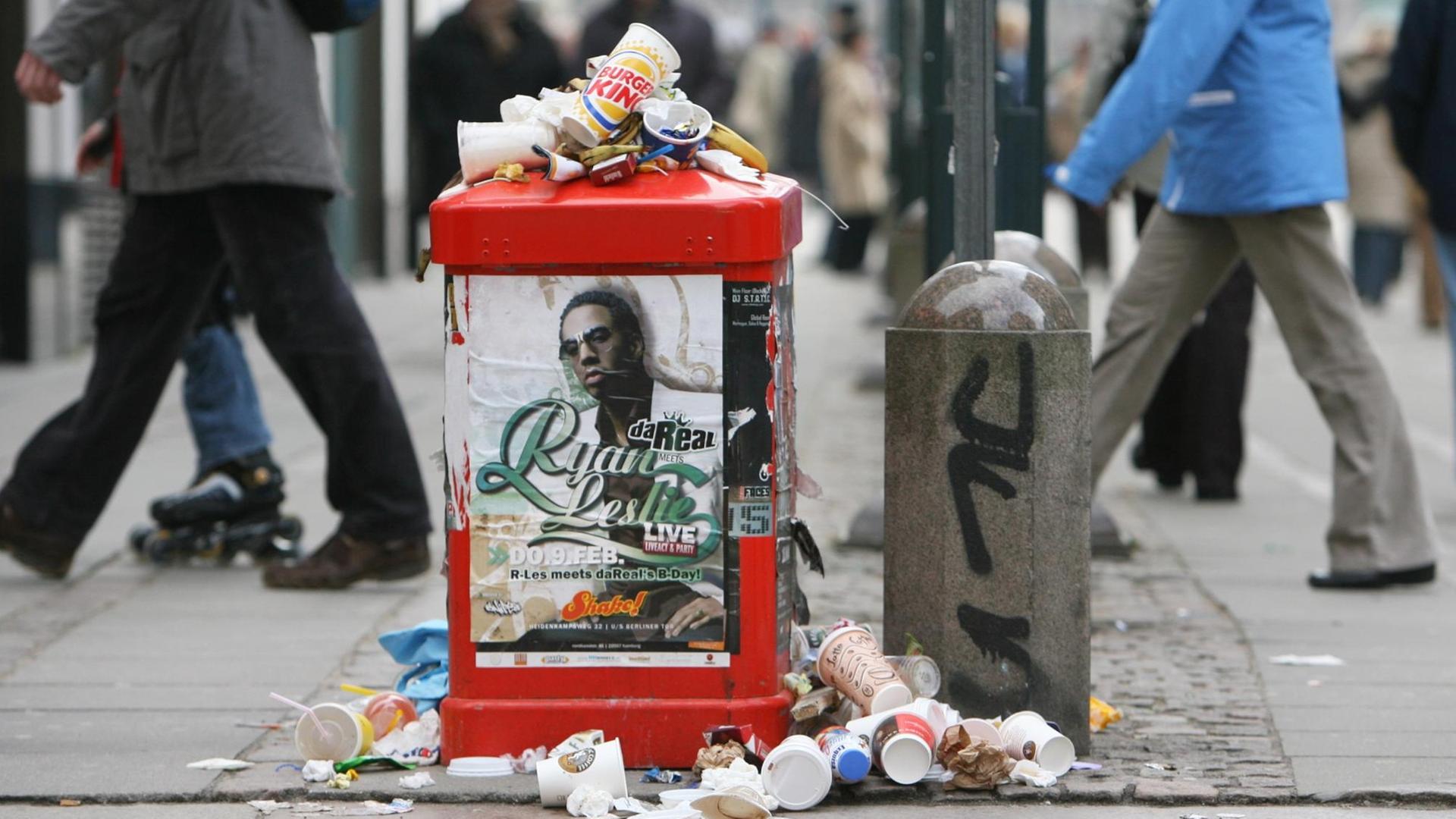 Passanten gehen in der Fußgängerzone in Hamburg an einem überfüllten Abfalleimer vorbei.